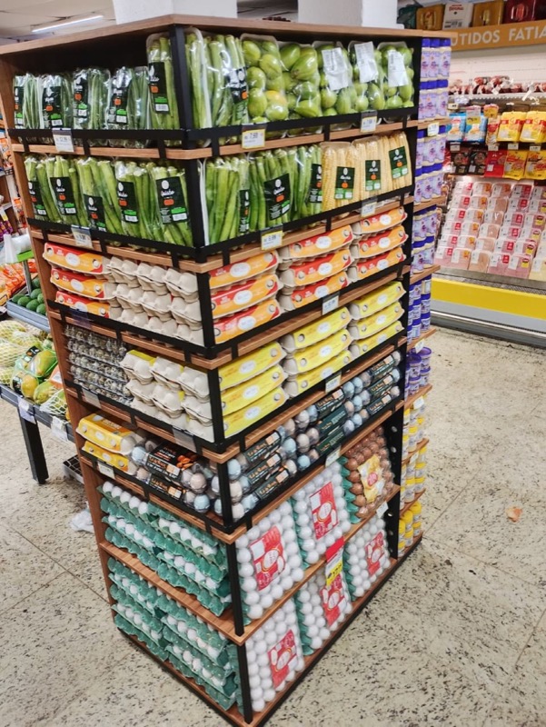 Expositor de dois lados carregado com ovos e legumes embalados, em aço preto com prateleiras em madeira.