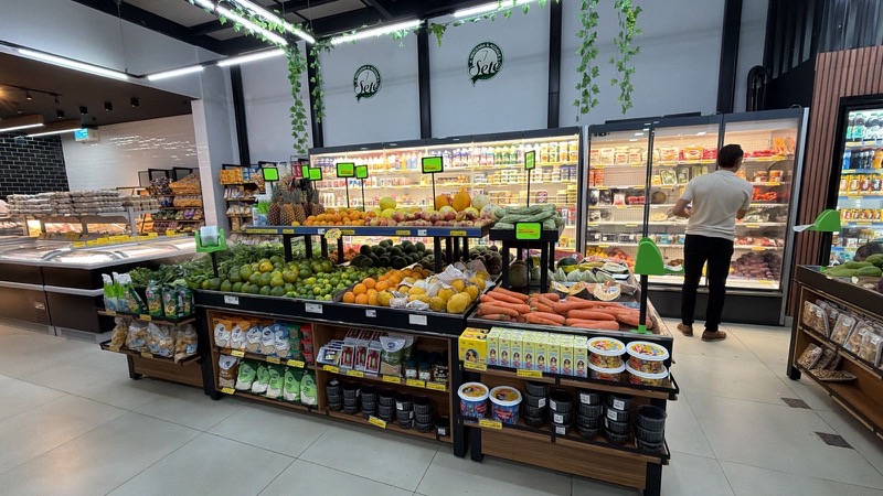 Ilha de hortifruti em padrão madeira com frutas e legumes, ao lado de expositores refrigerados.